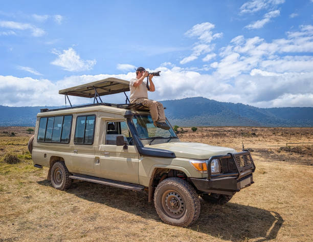 A wildlife photographer perched on the roof of a safari vehicle in Ngorongoro, Tanzania, captures the stunning scenery and wildlife through a telephoto lens. The rugged terrain, rolling hills, and expansive skies highlight the adventure and beauty of exploring Africa’s iconic landscapes and nature reserves.